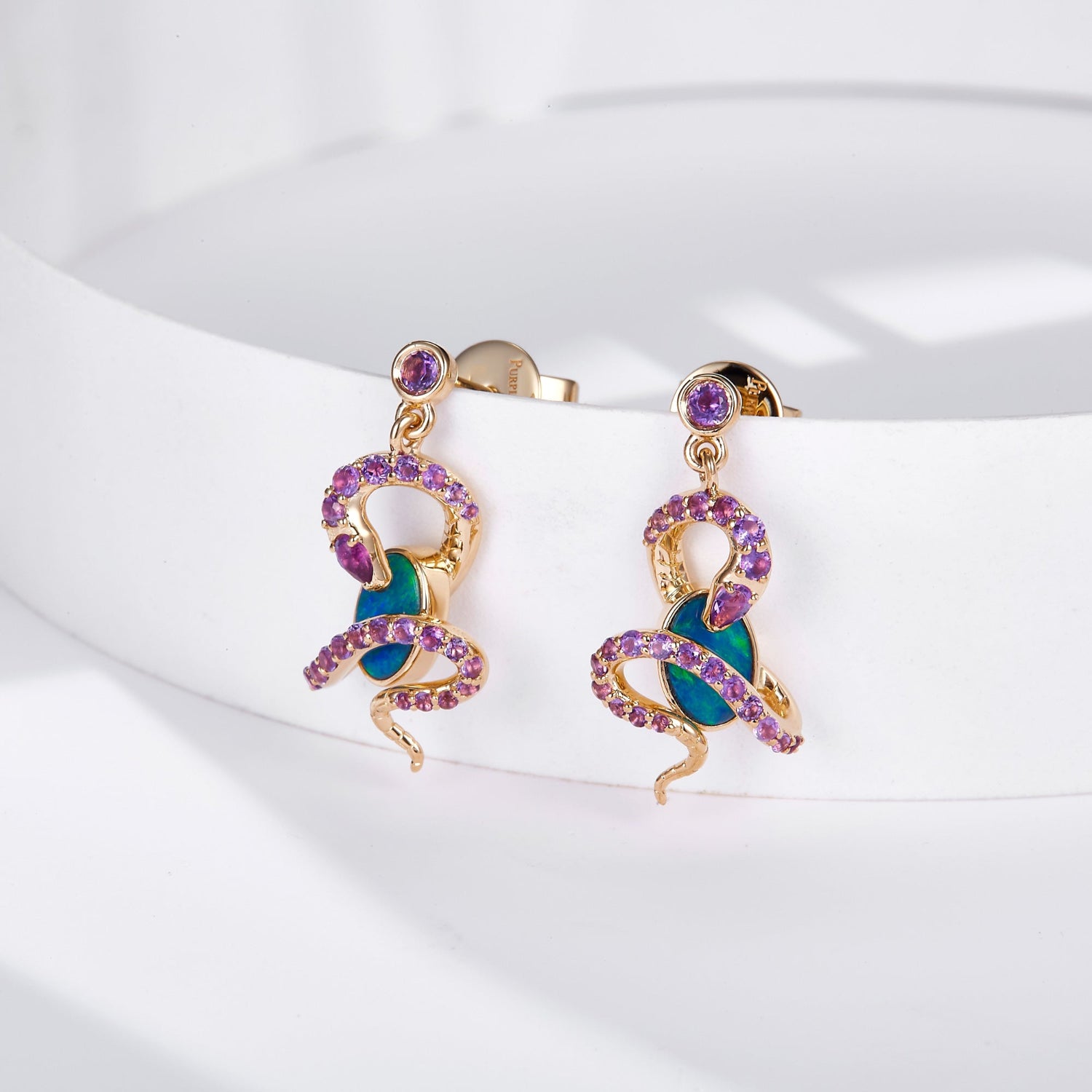 A pair of 18K gold dangle earrings featuring an intricate snake design wrapped around oval Australian opals, adorned with natural amethyst gemstones for a striking contrast. A round amethyst gemstone accents the stud, enhancing the luxurious appeal. The opals display vibrant blue-green hues, beautifully set in yellow gold. These statement earrings blend bold artistry with elegant sophistication, perfect for unique and luxurious styling.