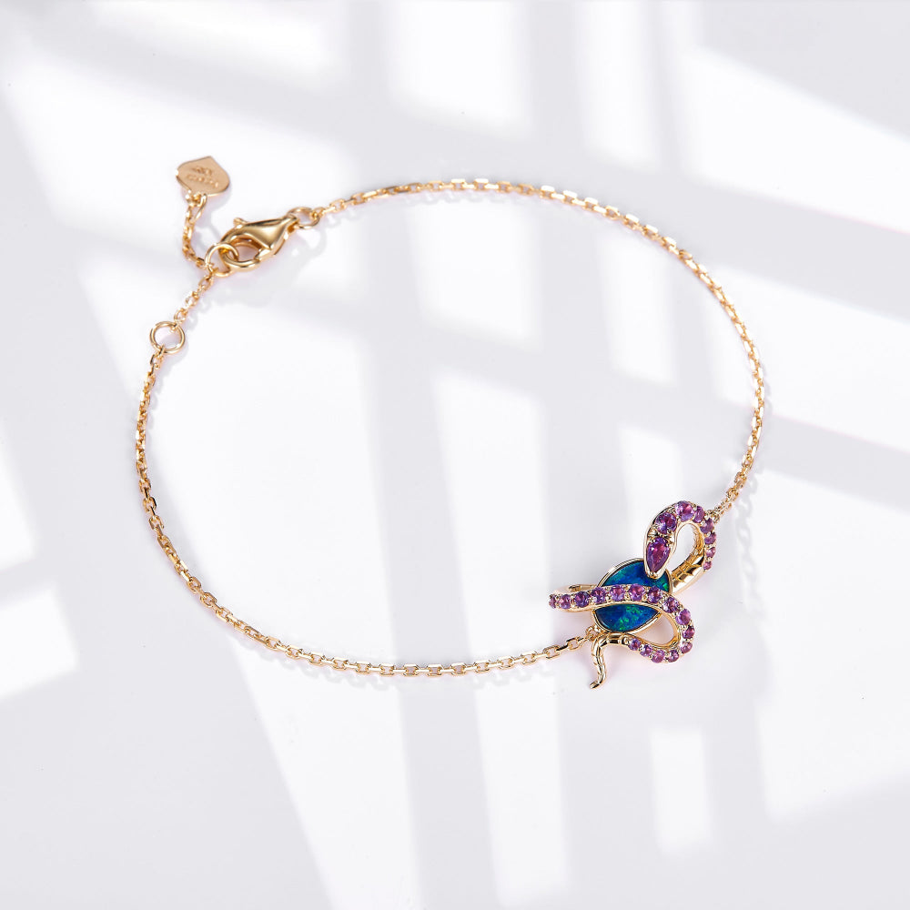 An elegant 18K gold bracelet featuring a snake design wrapped around an oval Australian opal. The snake is adorned with sparkling amethyst gemstones, complementing the vibrant blue-green hues of the opal. The delicate gold chain is secured with a lobster clasp and includes a small heart-shaped tag for a refined touch. This bracelet blends luxury with unique design, perfect for statement styling.